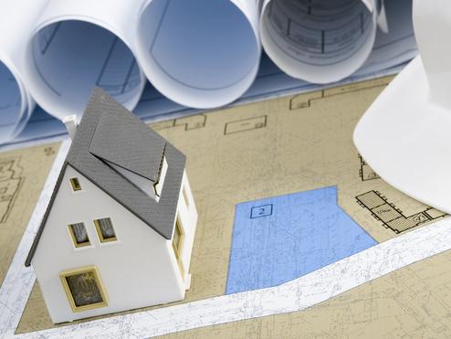 Modell eines Hauses auf Bauplänen, umgeben von einem Bauhelm und gerollten Plänen im Hintergrund.